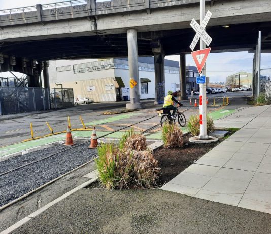 SDOT Adds Gravel Pit to Dangerous Burke-Gilman Track Crossing in Ballard A railroad crossing sign marks the spot next to some shrubs.
