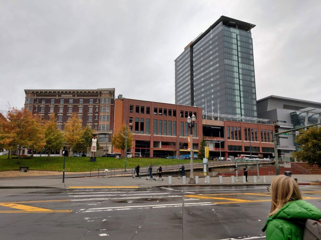 Four people on scooters zip across the plaza with a glass tower and older midrise brick buildings along Pacific Avenue in the background.