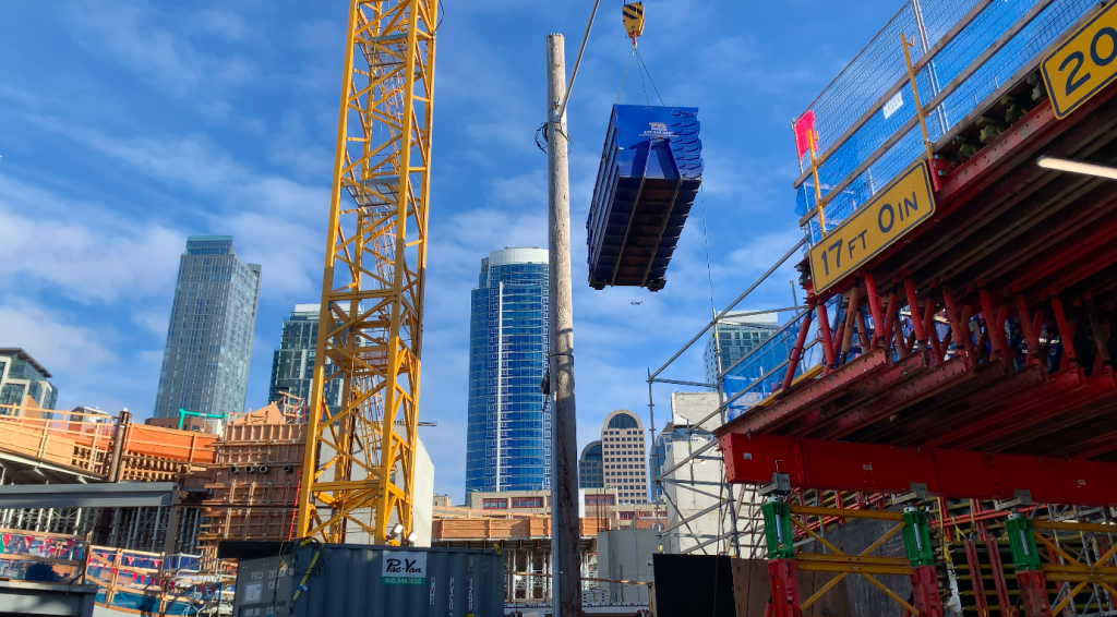 A blue container is moved by crane at a construction site.