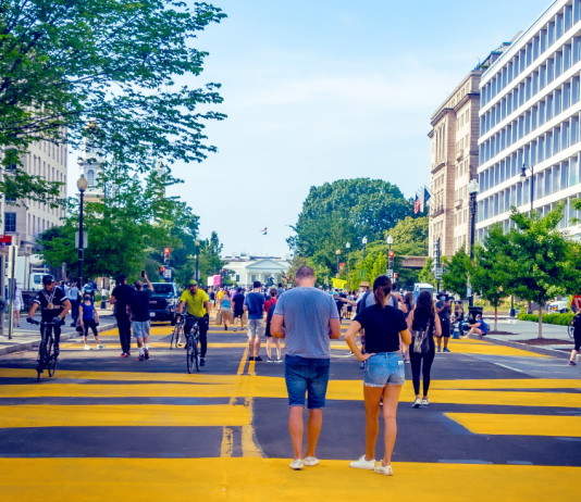 Report Ranking US Metros Shows Walkable Urbanism Pays Dividends People walk and bike on a wide street painted with yellow lines.