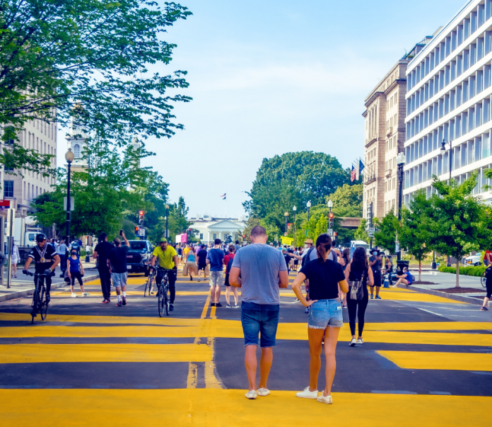 People walk and bike on a wide street painted with yellow lines.