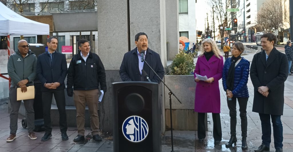 Mayor Bruce Harrell stands at a podium flanked by officials including Greg Spotts immediately to his right