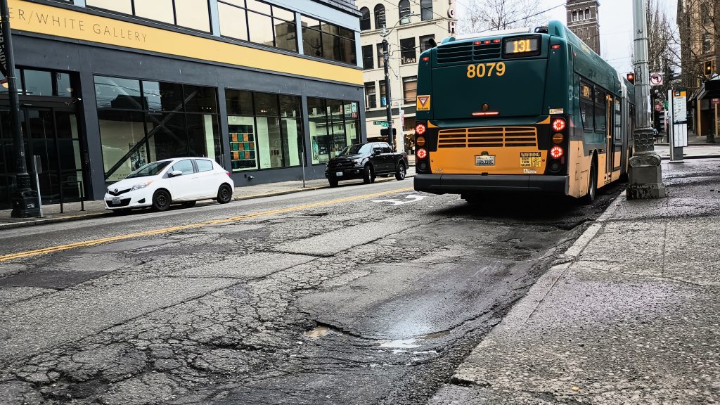 A closeup of the badly damaged pavement on 3rd Ave S with a bus waiting at the stop