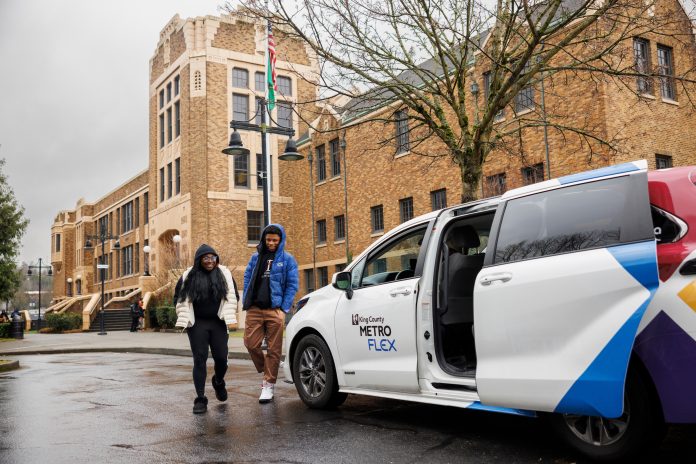 White van with passenger door open waits for riders in front of a brick building.