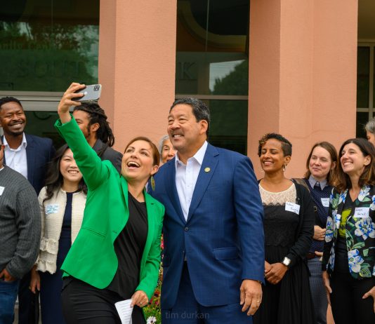 Mayor Harrell Unveils $970 Million Housing Levy Proposal Teresa Mosqueda reaches out her arm for a selfie with Mayor Bruce Harrell with several city staffers standing behind them.