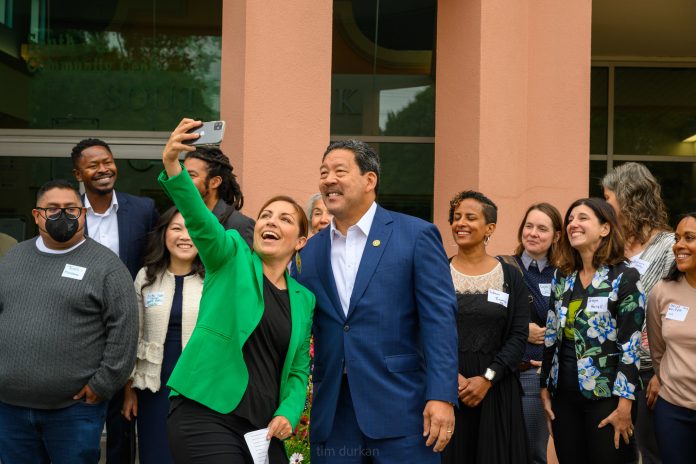 Mosqueda-and-Bruce-Harrell-selfie-Seattle-City-Council-flickr Teresa Mosqueda reaches out her arm for a selfie with Mayor Bruce Harrell with several city staffers standing behind them.