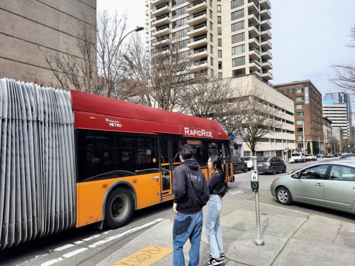 RapidRide-H-in-Belltown-with-peds-20230319_144837 Two pedestrians and a car watch the RapidRide H bus go by.