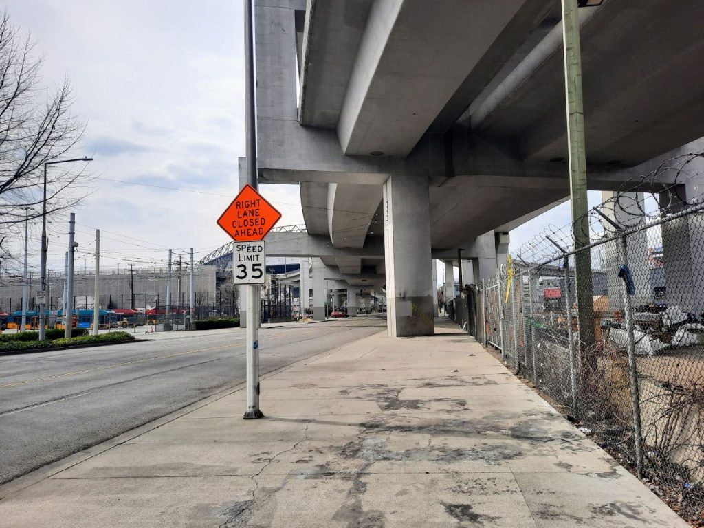 A pockmarked sidewalks next to a razorwire fence underneath a I-90 freeway overpass is the pedestrian experience people are offered on South Royal Brougham Way.