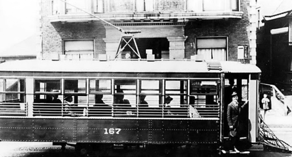 Black and white image of a street car numbered 167 running in the middle of a street in front of a two story house.