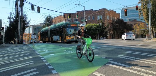 Someone riding a lime bike across a bike lane in Capitol Hill with a bus behind them