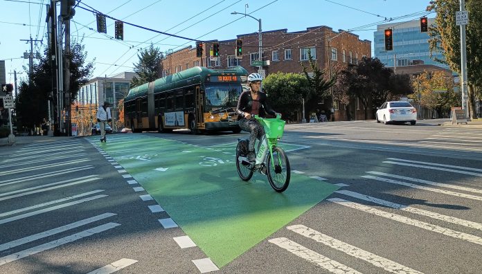 Someone riding a lime bike across a bike lane in Capitol Hill with a bus behind them