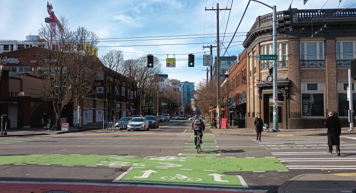 Someone on a bike crosses the street as pedestrians walk in a crosswalk and drivers wait to turn at Broadway and Pike in Seattle