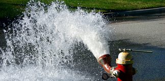 A red fire hydrant shows a five foot geyser of water on some green grass.