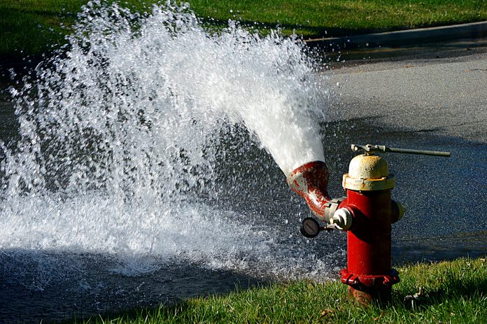 A red fire hydrant shows a five foot geyser of water on some green grass.