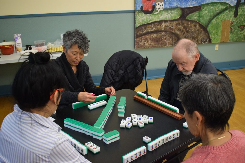 four adults sit at a table and play a game with tiles