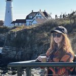 Erin Caldwell, long red hair, ball cap, standing in front of a lighthouse