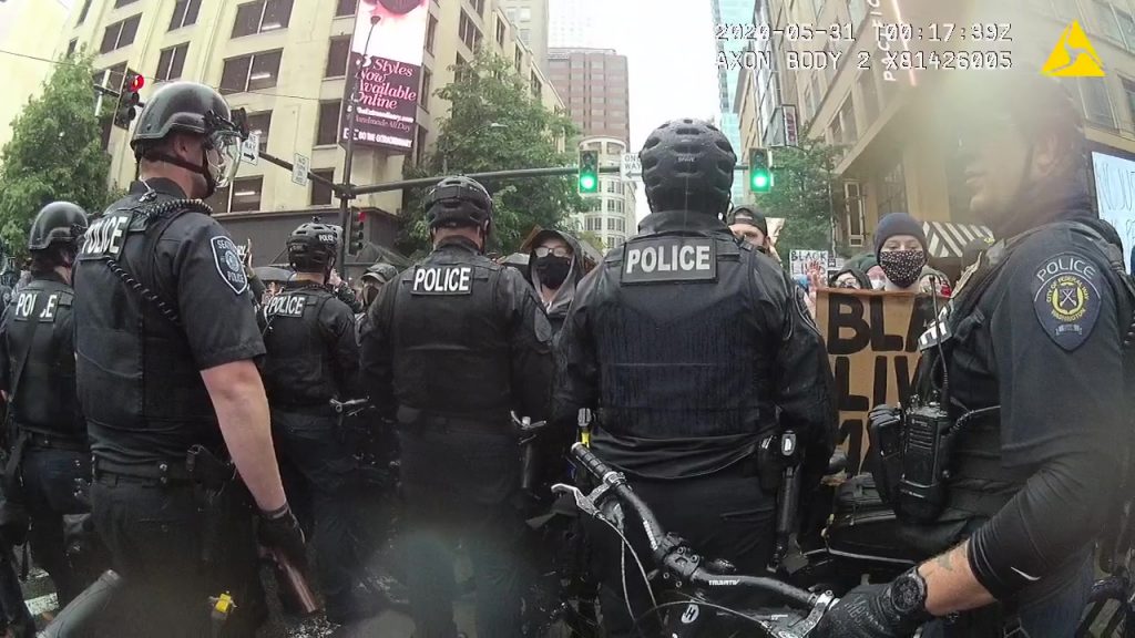 A line of police offers in riot gear face off against protesters sporting Black Lives Matter signs along a Downtown Seattle street.