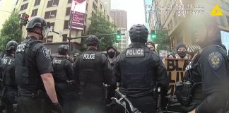 A line of police offers in riot gear face off against protesters sporting Black Lives Matter signs along a Downtown Seattle street.