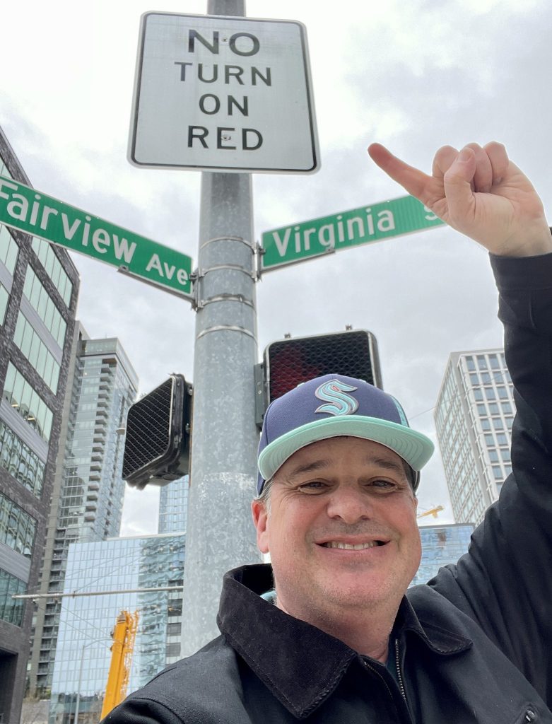 A man pointing at two street signs, one of which says Fairview Ave and the other says  Virginia Street, with a no turn on red sign between them
