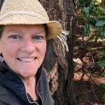 Linda Hanlon next to a tree and rhododendron bush.