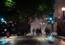 Chaos and Banned Weapons: SPD’s Use of ‘Mutual Aid’ Police Departments in 2020 In a night time image, dark police officer silhouettes emerge from an armored truck amidst a backdrop of a cloud of tear gas.