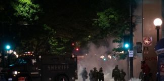 In a night time image, dark police officer silhouettes emerge from an armored truck amidst a backdrop of a cloud of tear gas.