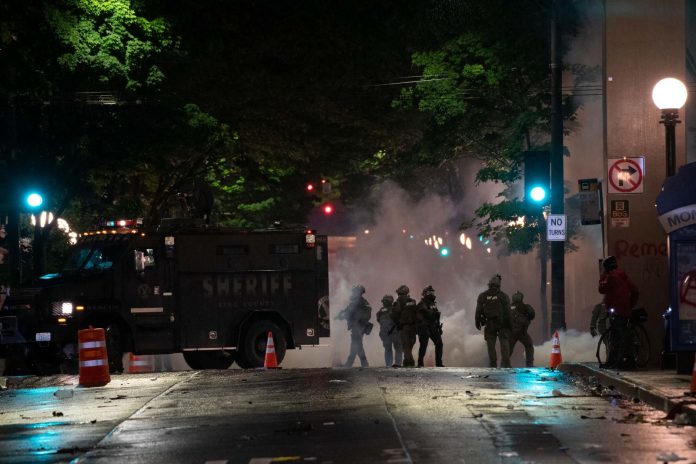 In a night time image, dark police officer silhouettes emerge from an armored truck amidst a backdrop of a cloud of tear gas.