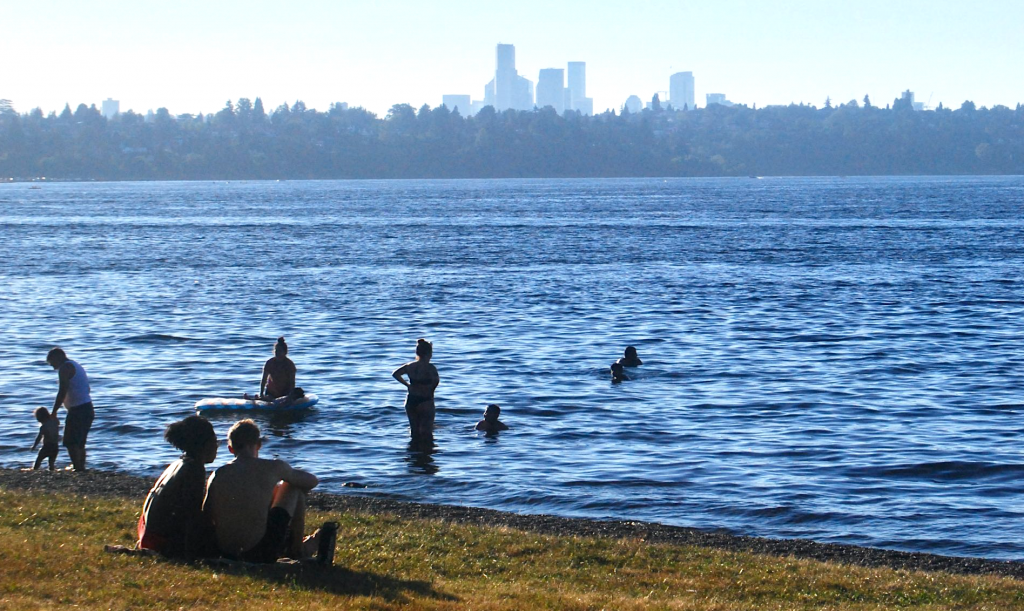 A half dozen people wade in the water at a Lake Washington beach with the Seattle skyline visible over the ridge in the background.