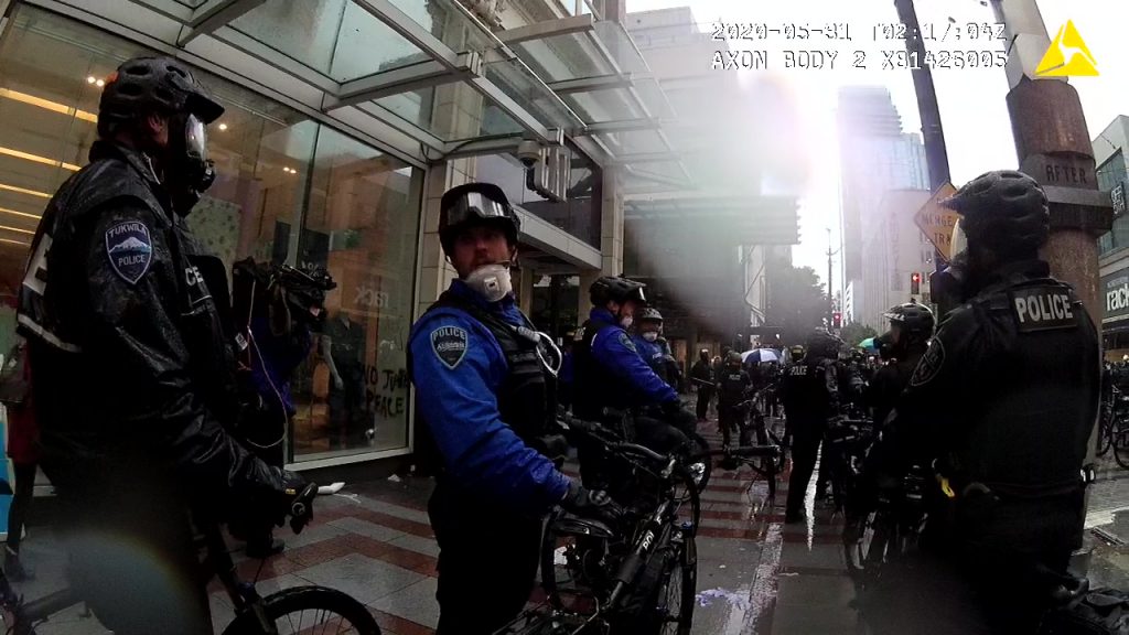 A rain-smudged body cam image shows a gaggle of police stand under an awning.
