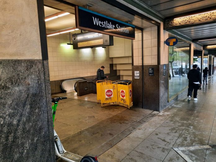 People walk by the station entrance, which includes a yellow barricade for a closed elevator.