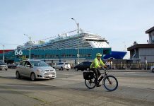 Are Cruise Ships Crushing Seattle’s Climate Goals? Bicyclist in neon jacket and helmet on busy street. Cruise terminal and ship behind him.