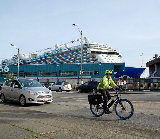Are Cruise Ships Crushing Seattle’s Climate Goals? Bicyclist in neon jacket and helmet on busy street. Cruise terminal and ship behind him.