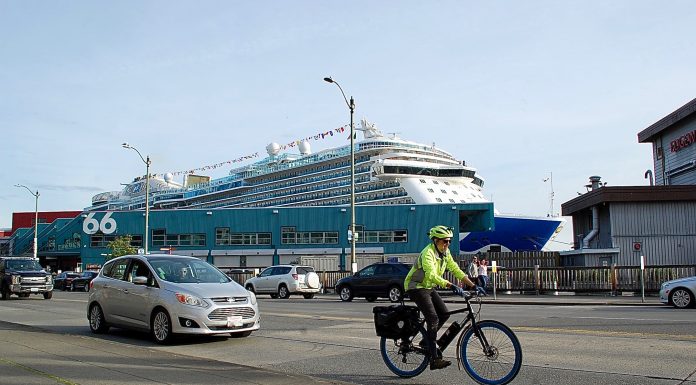 Are Cruise Ships Crushing Seattle’s Climate Goals? Bicyclist in neon jacket and helmet on busy street. Cruise terminal and ship behind him.