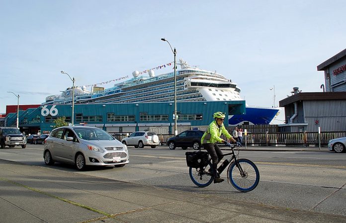 Bicyclist in neon jacket and helmet on busy street. Cruise terminal and ship behind him.