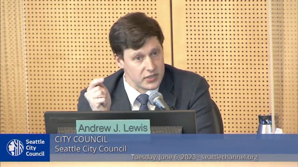 Andrew Lewis wears a suit and gestured with a crooked finger at the dais in Council Chambers.