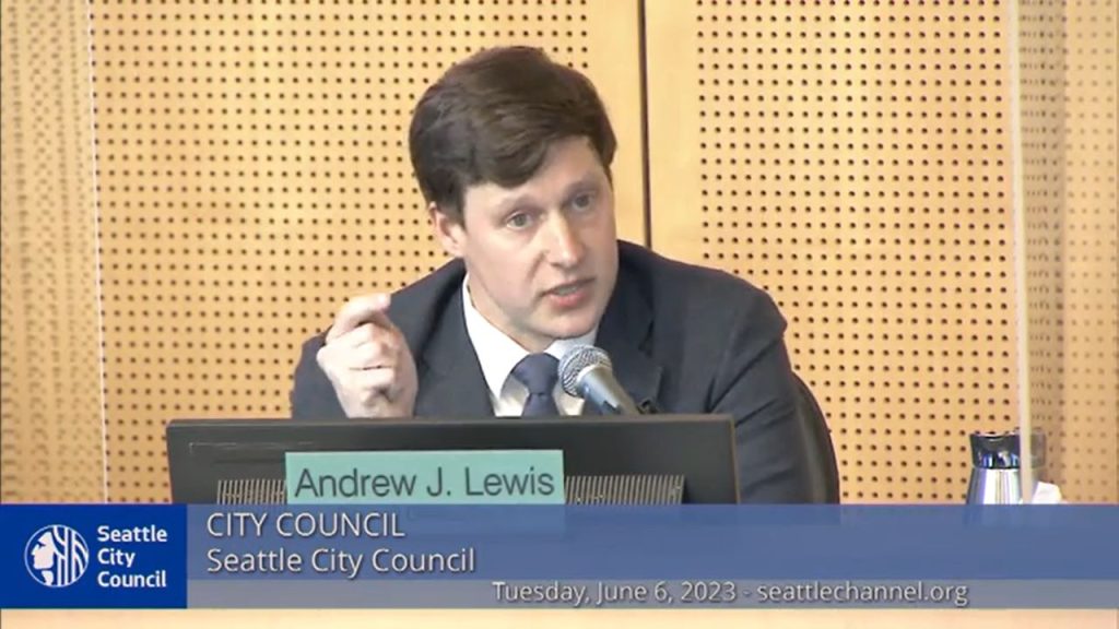 Andrew Lewis wears a suit and gestured with a crooked finger at the dais in Council Chambers.