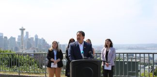 Bruce Harrell in a suit standing at a lectern. MLK Labor’s Executive Secretary-Treasurer Katie Garrow is to the left and OSE Director Jessyn Farrell to the right. A hazy Seattle skyline is in the background from Queen Anne vantage.