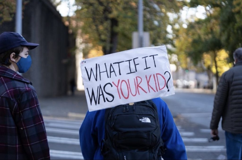 A sign reads "What if it was your kids?" as three protesters cross a crosswalk.