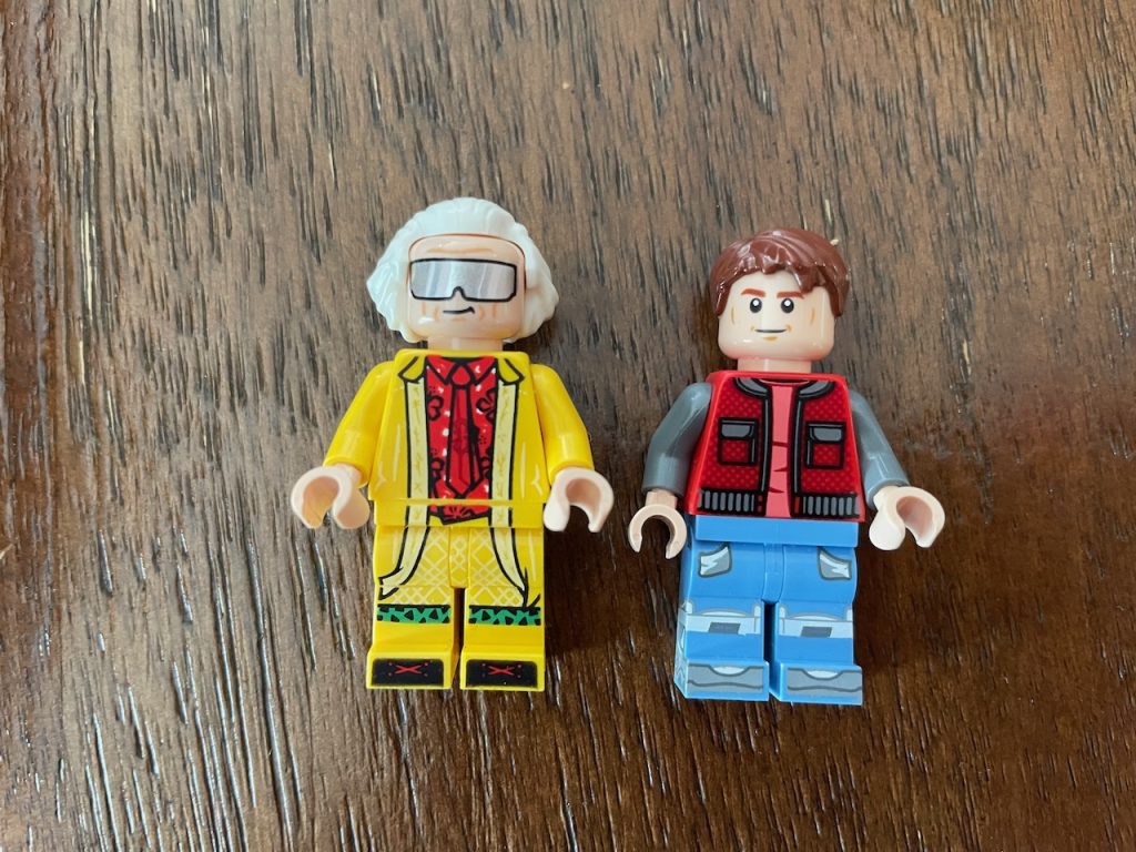 Two Lego minifigs, left is in yellow with white hair and wrap around sunglasses. Right is in a vest with a smirk and brown hair.