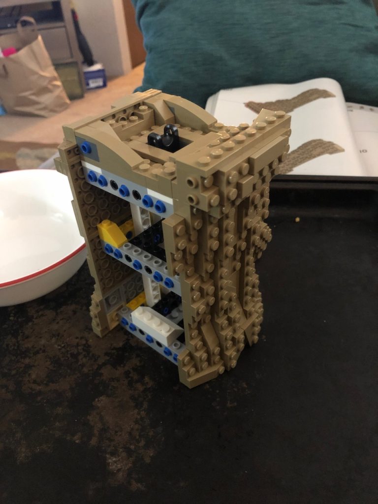 Two brown bumpy lego surfaces attached to the sides of a lego tower to form the base of a Grogu statue.