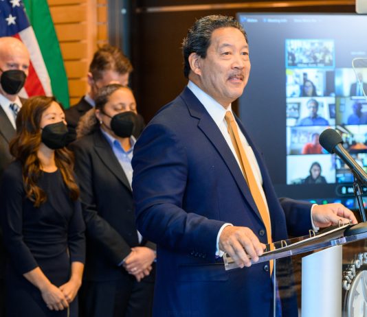 Is Harrell Drifting to the Right Amidst Deputy Mayor Shakeup? Harrell stands at a lectern with a grin. His deputy mayors and Burgess wear masks.