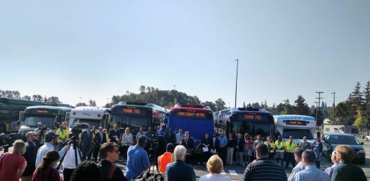 King County Executive Dow Constantine speaks at a podium in front of a row of busses flanked by Metro employees.