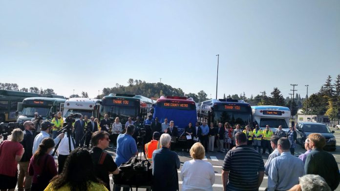 King County Executive Dow Constantine speaks at a podium in front of a row of busses flanked by Metro employees.