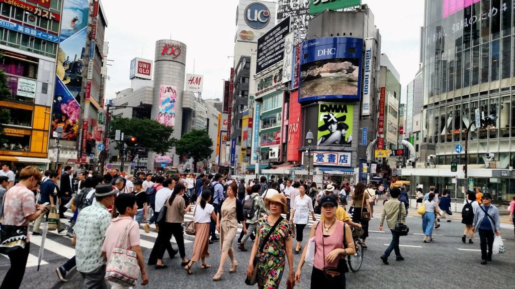 More than 100 people cross an intersection in the bustling Shibuya neighborhood of Tokyo. Ads for shops dot the buildings.