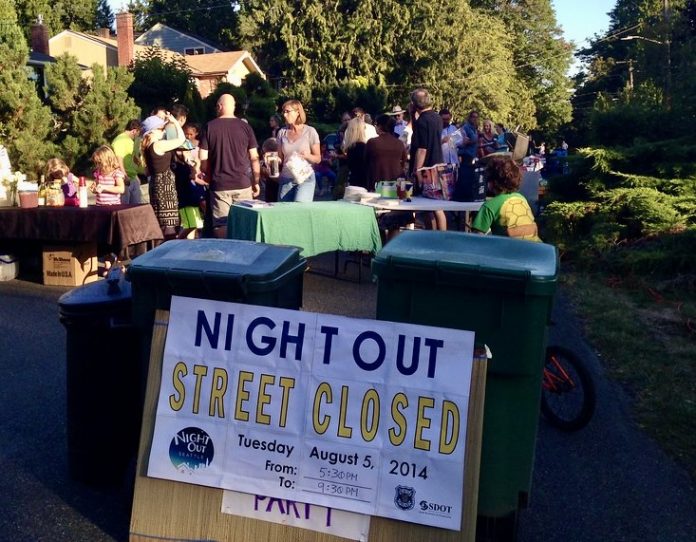 A sign reads Night Out street closed with people gathered behind it socializing.