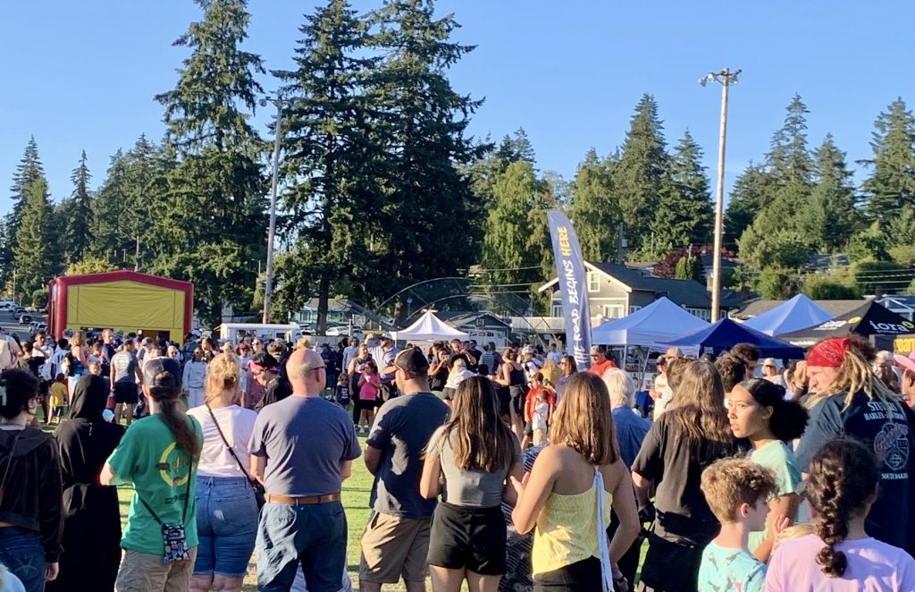 A crowd of people gather near booths and tall trees.