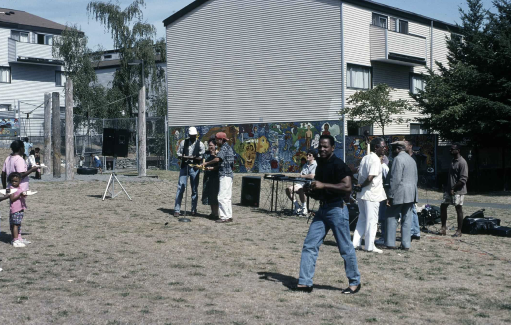 People play musical instruments and gather in a park next to a large building with a mural. 
