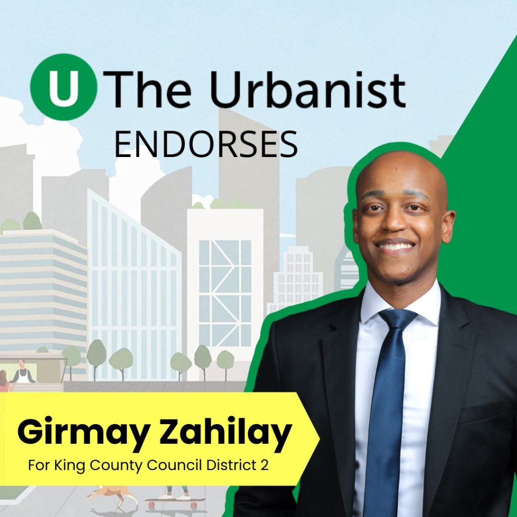 Girmay is a Black man in a dark suit and smiles in this campaign photo. The graphic has a city skyline as the background with The Urbanist logo.