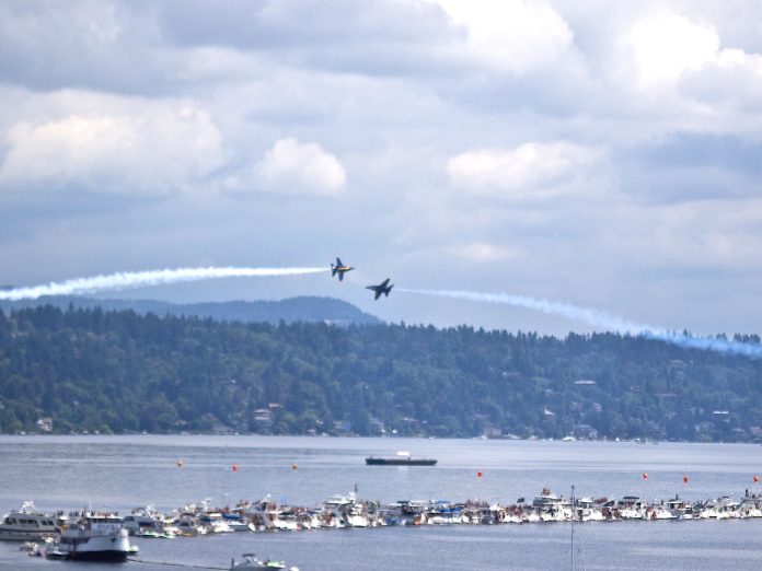Two Navy Blue Angels planes pass each other over boats tied to a log boom in Lake Washington.