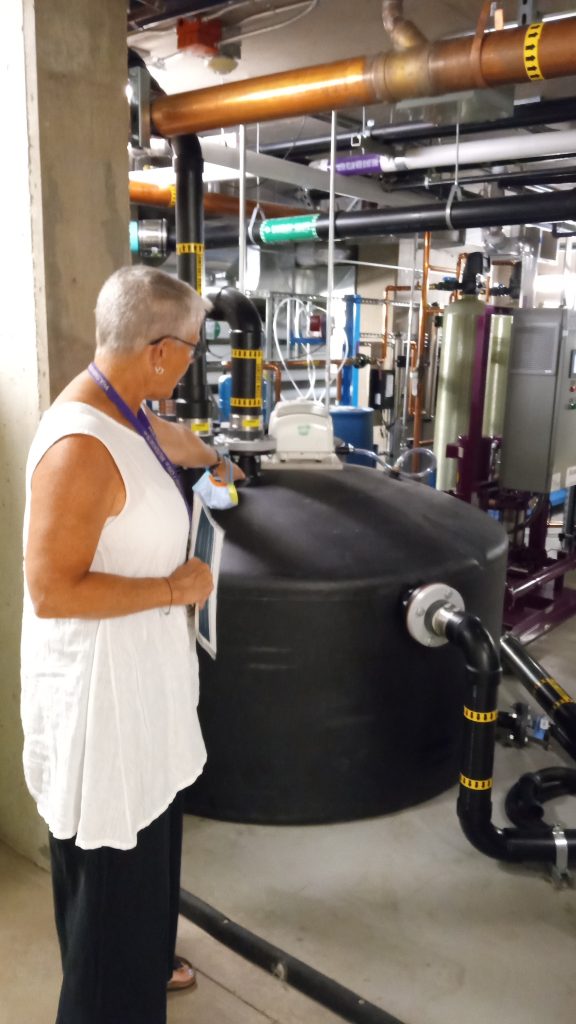 A women with short hair, white top, black pants points to a black tank that collects gray water for recycling.